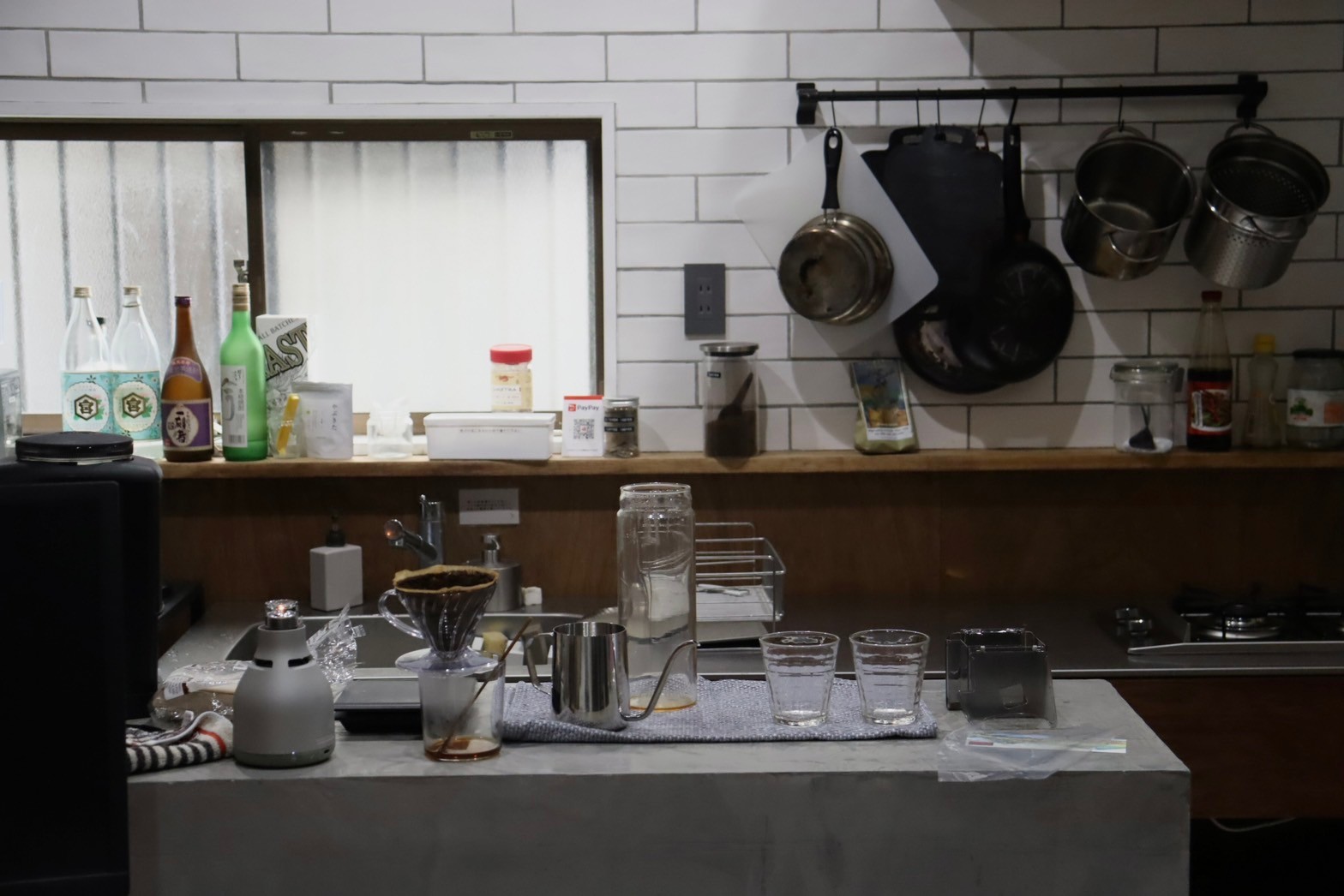 キッチンを正面から撮った写真。右手背景にはフライパンや鍋が引っ掛けられていて、カウンターには酒の瓶や調味料が並んでいる。手前にはコーヒーを淹れるための器具、ポット、たくさんのグラスが机に置いてある。